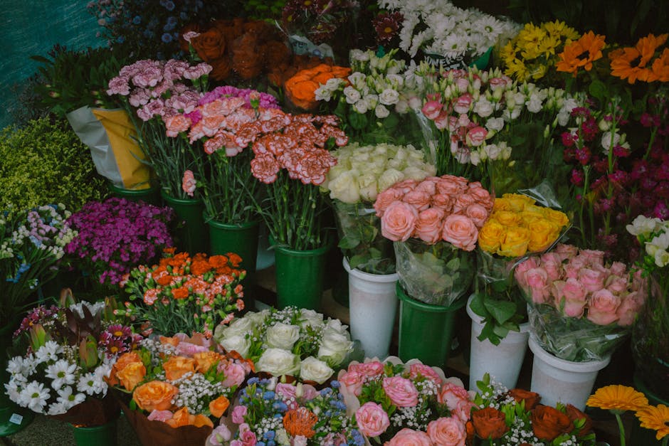 A vibrant display of fresh floral bouquets and potted plants arranged on tables and shelves at a flower shop, featuring a variety of flowers in shades of pink, white, yellow, orange, and purple. The b